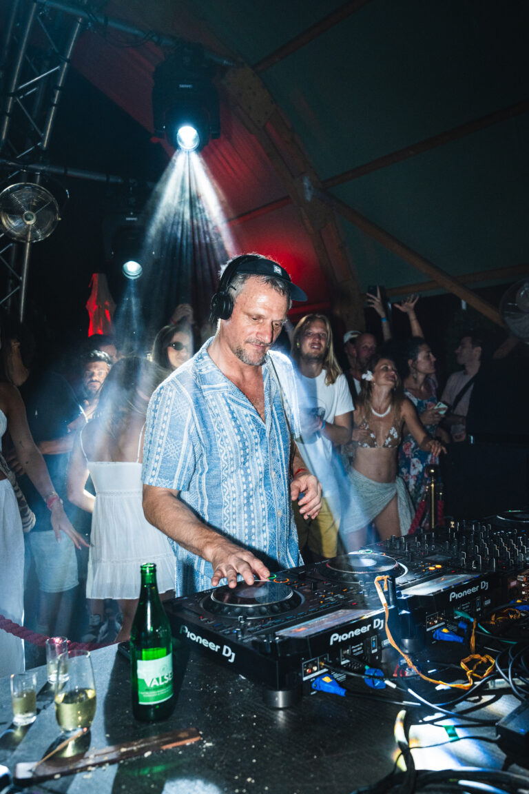 Ein Mann mit Kopfhörern bedient ein Pioneer-DJ-Pult in einer gut gefüllten abendlichen Location unter hellem Bühnenlicht.