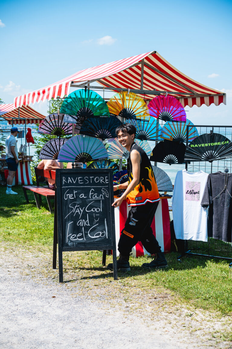 Ein lächelnder Mann steht hinter einem Marktstand im Freien, der bunte Fächer und eine Tafel mit der Aufschrift RAVESTORE Get a fan Stay Cool and Feel Cool präsentiert.