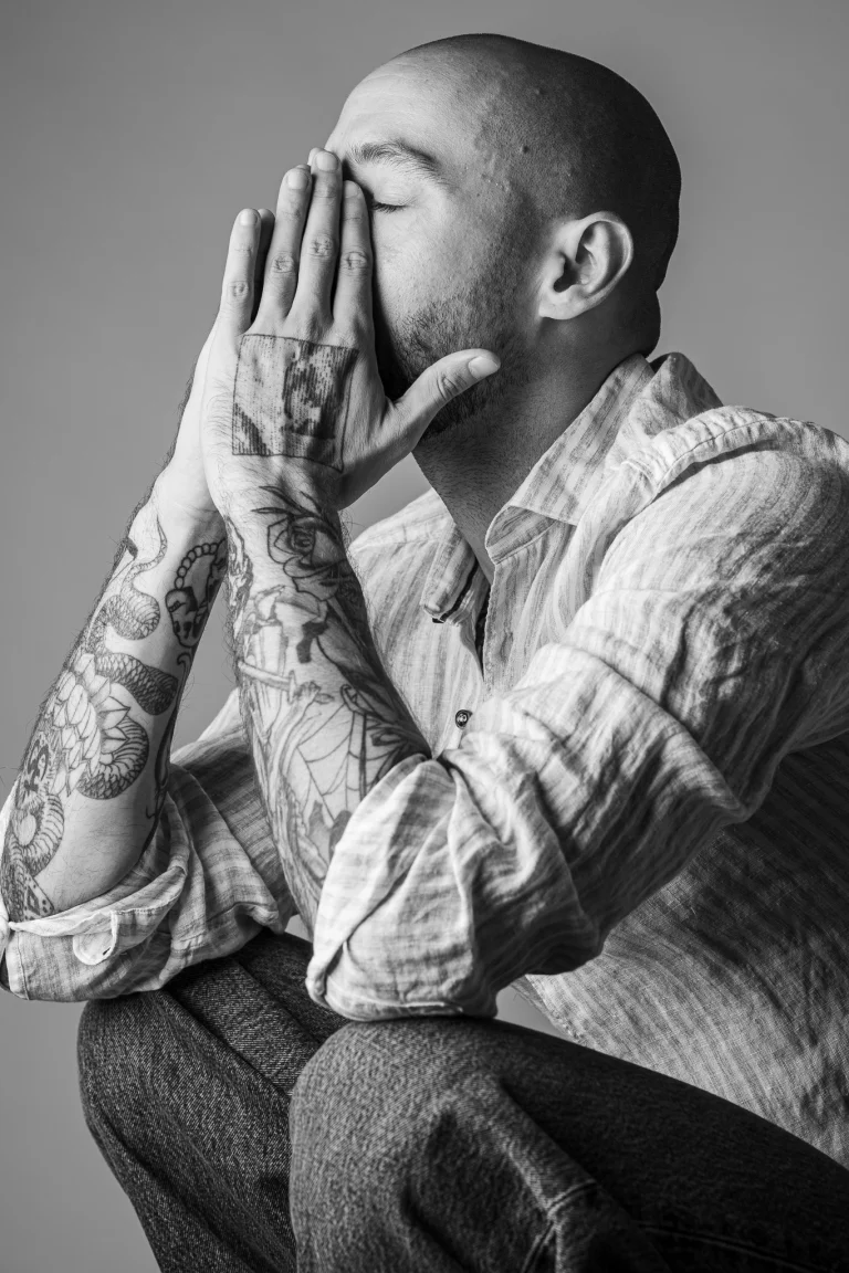 A black and white studio portrait of a bald tattooed man crouching with his face covered by his hands.