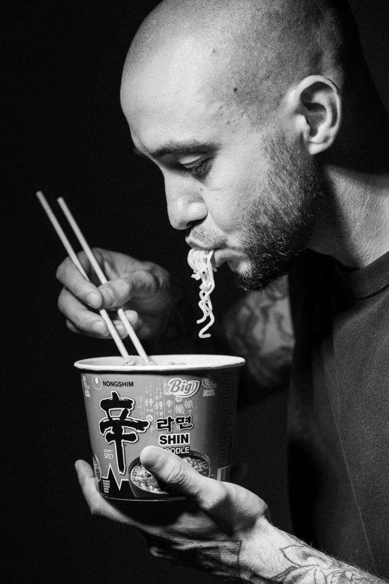 A bald man with a beard eats noodles with chopsticks from a paper cup in a black and white close-up.
