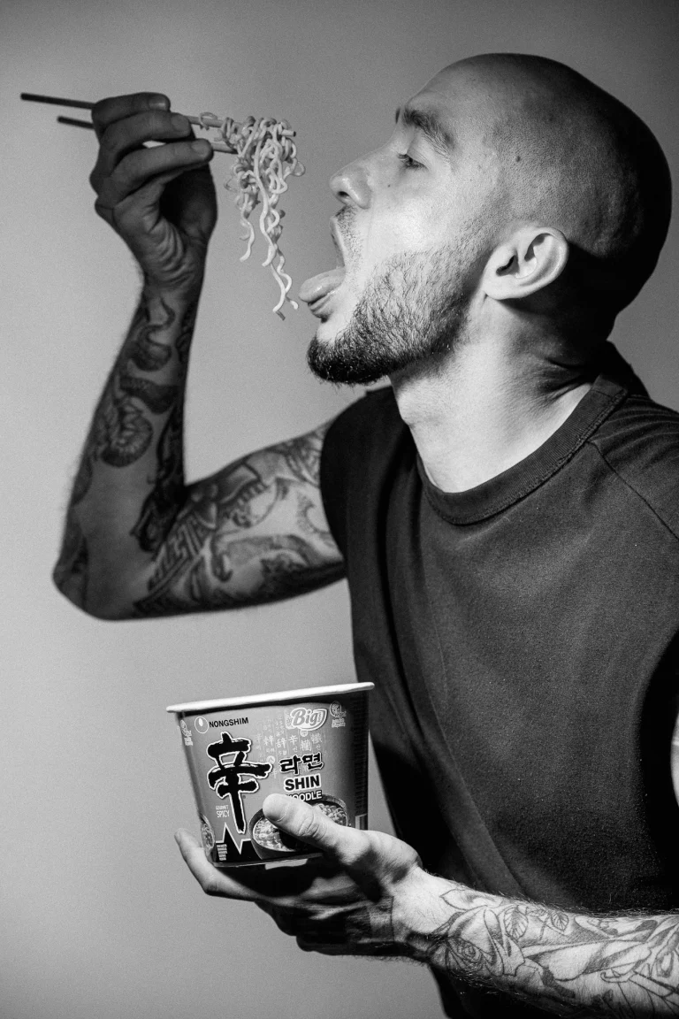Black and white portrait of a tattooed man holding a cup of Shin noodles, using chopsticks to lift noodles toward his open mouth.