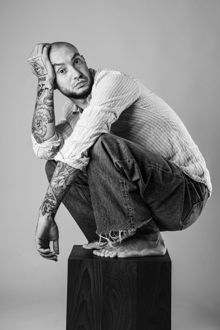 A bald man with heavily tattooed arms crouching barefoot on a wooden block against a plain backdrop.