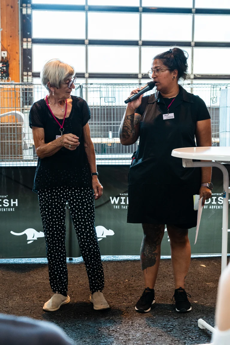 An older woman in a black shirt and polka-dot trousers stands talking to another woman holding a microphone indoors.