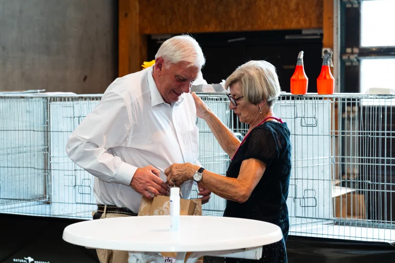 A man in a white shirt and a woman in a black top look down at a small bag indoors near wire animal cages.