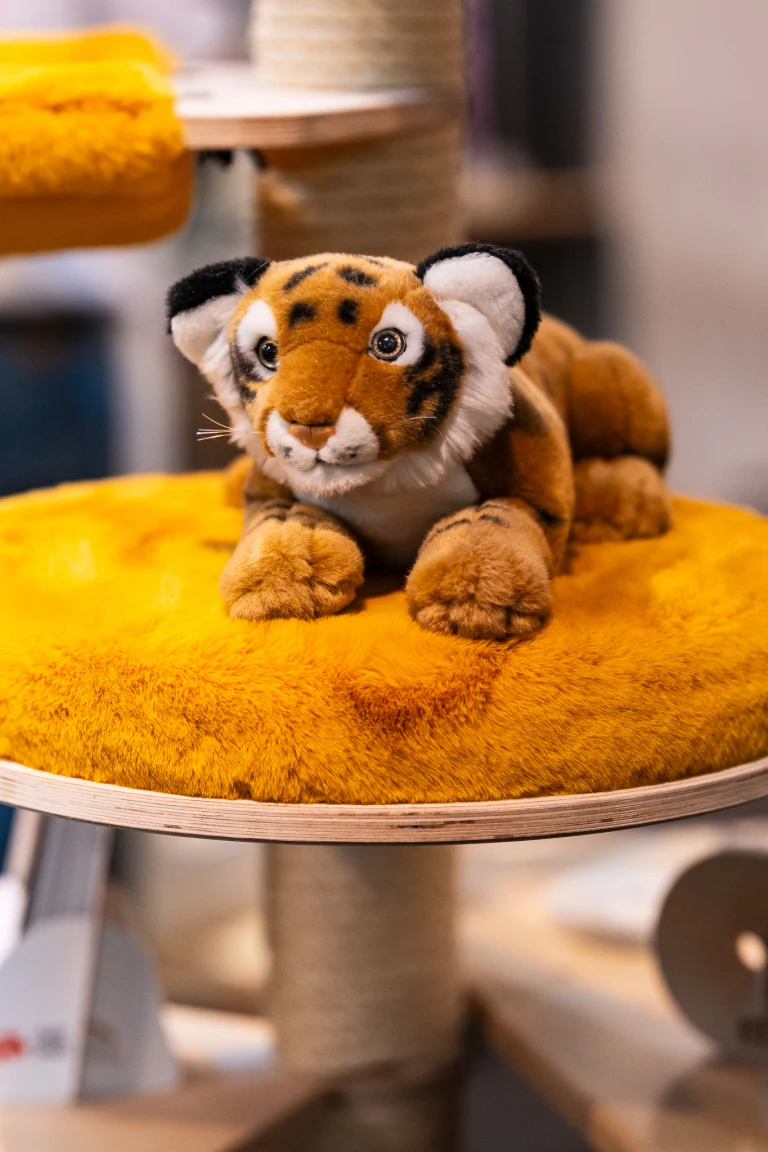 A plush toy tiger lies on a round, yellow fuzzy platform of a cat tree.