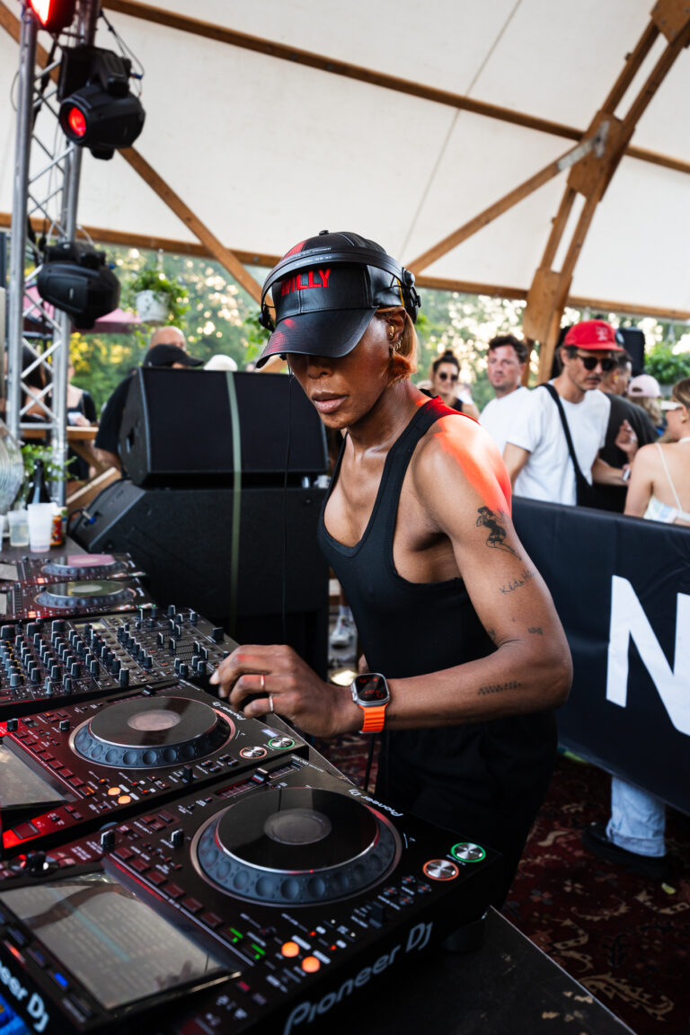 Eine Frau mit schwarzer Kappe und Trägershirt bedient ein DJ-Mischpult mit Plattentellern.