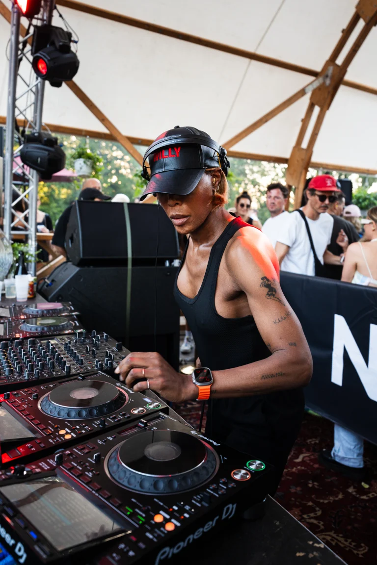 A woman wearing a black cap and tank top operates a DJ mixer with turntables.
