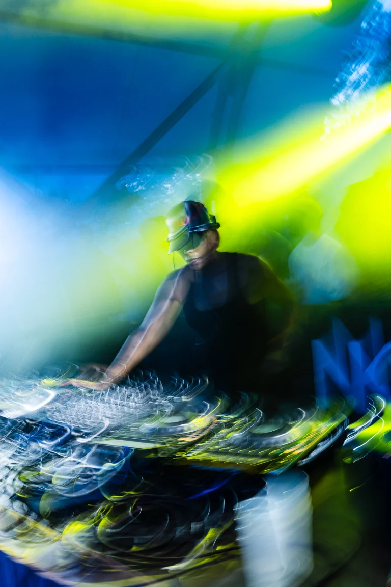 A woman wearing a cap mixing music on a DJ deck with motion blur under blue and yellow stage lighting.