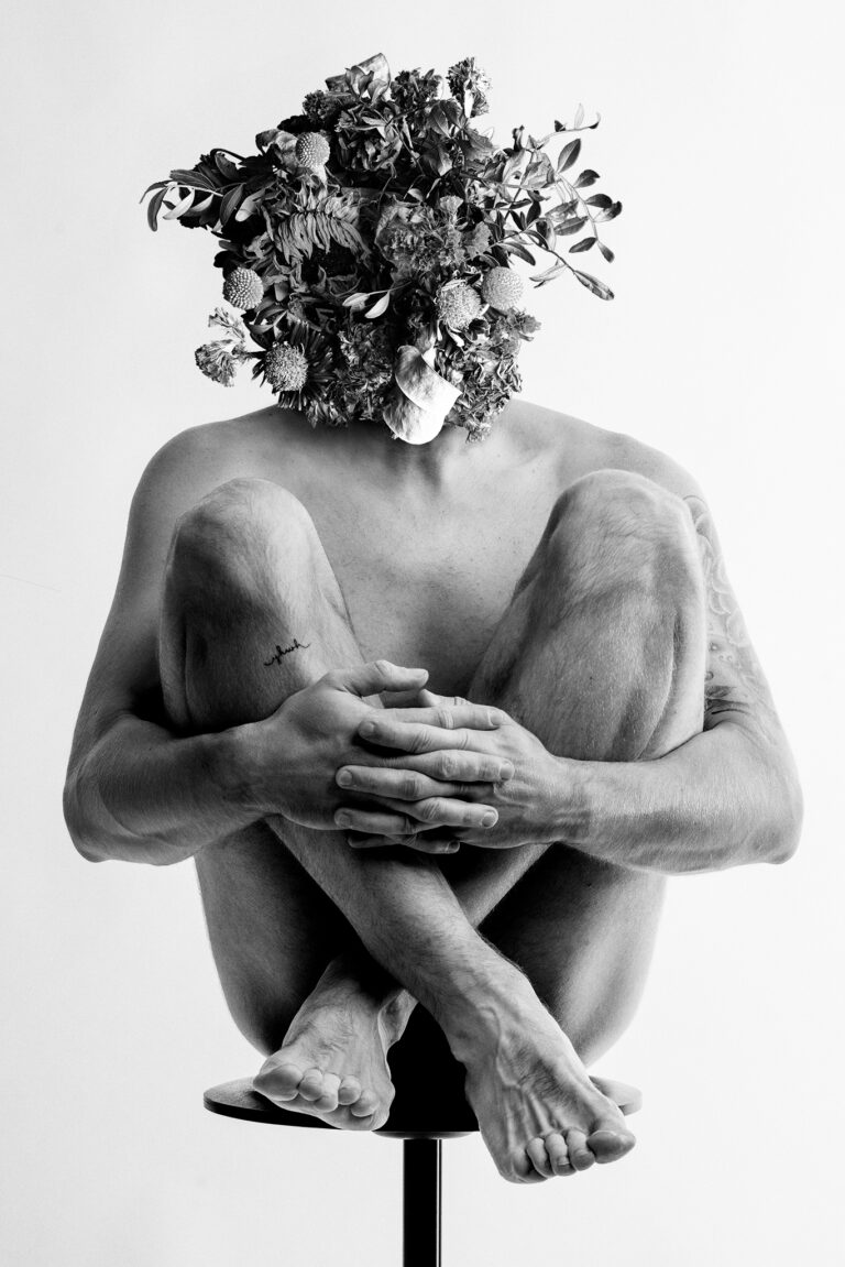 A black and white studio portrait of a seated man with a large bouquet of dried flowers completely obscuring his head.