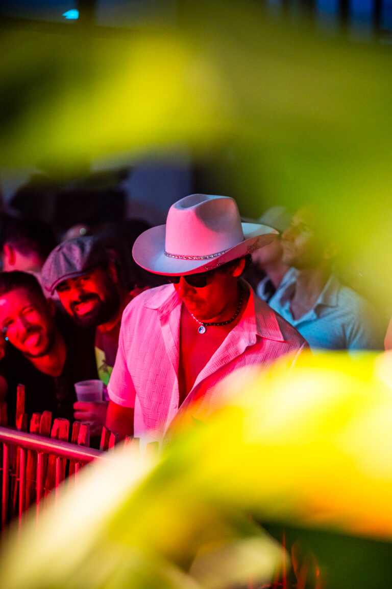 Ein Mann mit weißem Cowboyhut, Sonnenbrille und hellem Hemd steht in einer dunkel beleuchteten Menge, die von rotem und gelbem Licht erhellt wird.