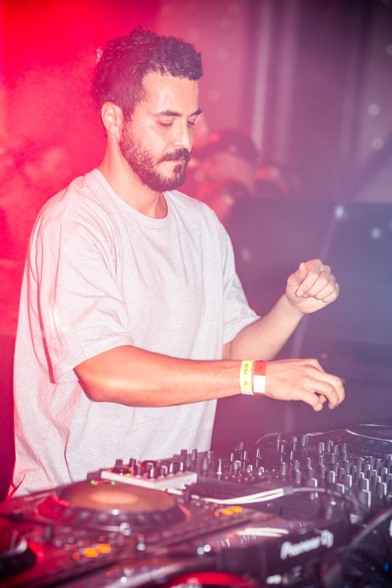 Ein bärtiger Mann in einem hellgrauen T-Shirt steht hinter einem DJ-Mixer und bedient die Regler unter rotem Bühnenlicht.