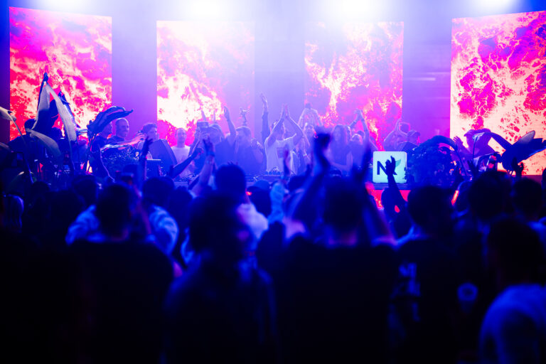 A crowd of people with raised hands facing a stage illuminated by bright pink screens under purple stage lights.