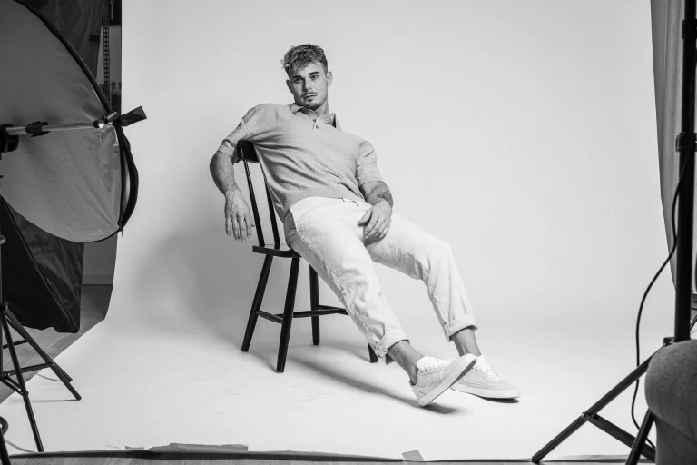 A black and white portrait of a man leaning back on a dark wooden chair in a studio with light equipment visible.