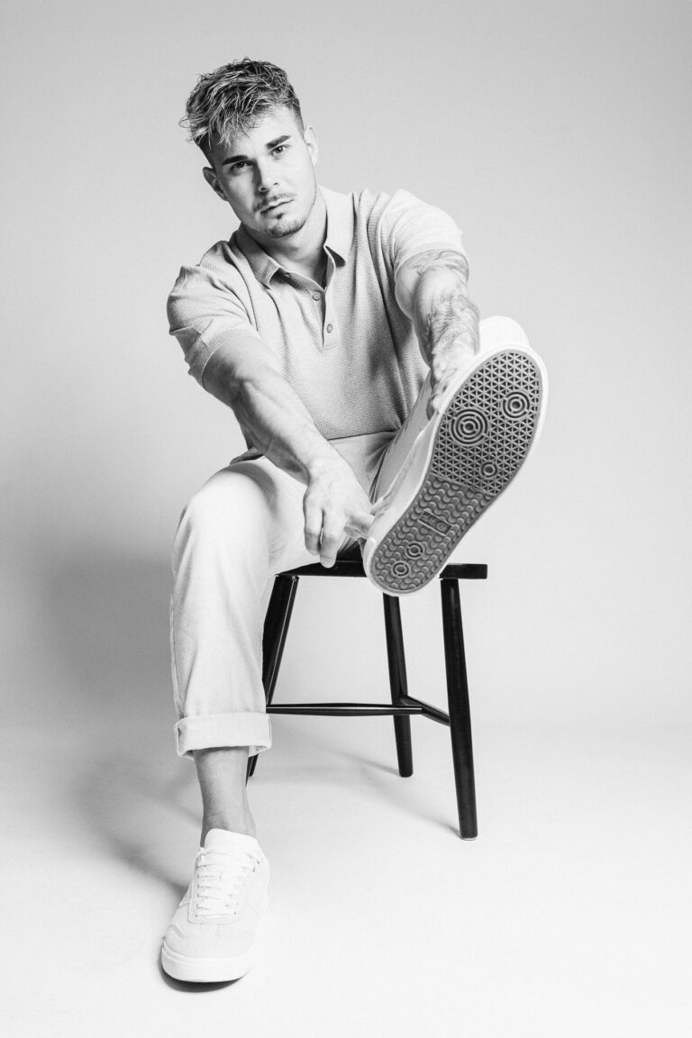 Black and white portrait of a man sitting on a stool and holding the sole of his sneaker toward the camera.