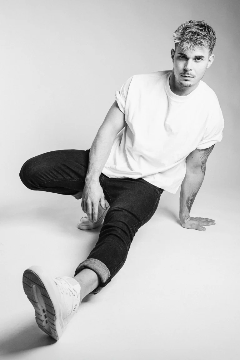 A black-and-white studio portrait of a man squatting against a plain background, wearing a white t-shirt, dark jeans, and sneakers.