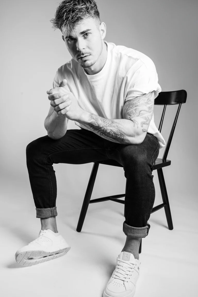 A man with tattoos sits on a dark chair against a light background, leaning forward with clasped hands.