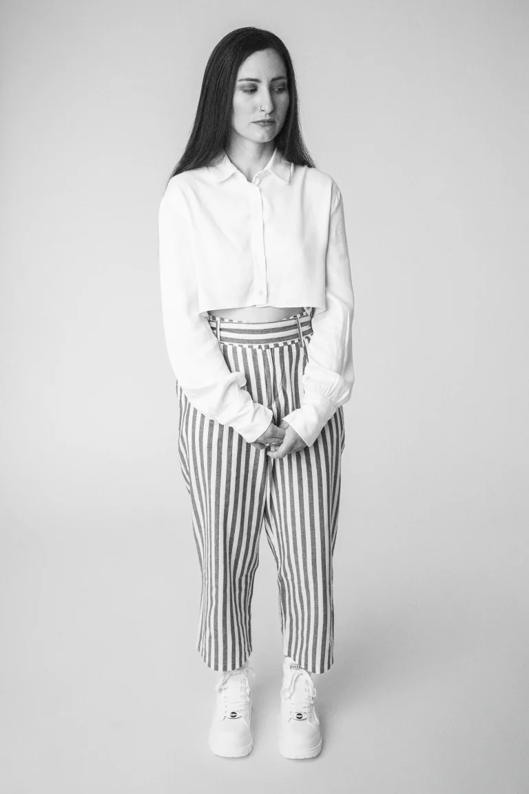 A black-and-white full-length studio portrait of a woman standing and looking down, wearing a cropped white button-down shirt, striped trousers, and platform sneakers.