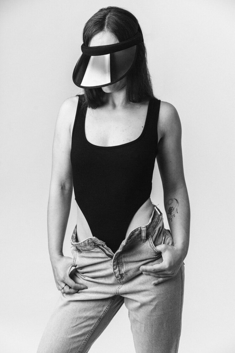 A black-and-white studio portrait of a woman wearing a black visor that covers her eyes, a black bodysuit, and unbuttoned light jeans.