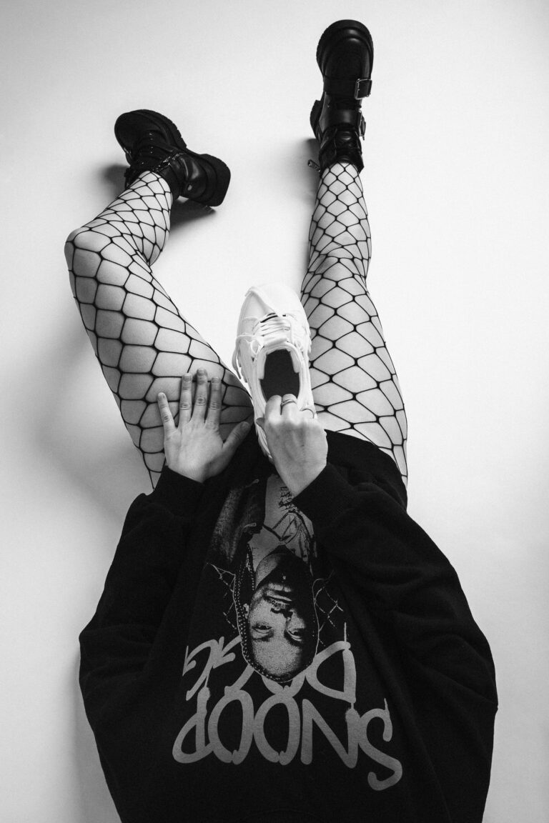 High-angle monochrome studio portrait of a woman sitting on the floor in fishnet tights and a black sweatshirt, holding a white sneaker.