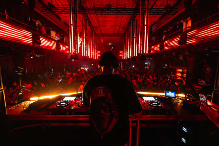 Ein Mann steht mit dem Rücken zur Kamera hinter einem DJ-Pult, beleuchtet von rotem Bühnenlicht in einem vollen Club.