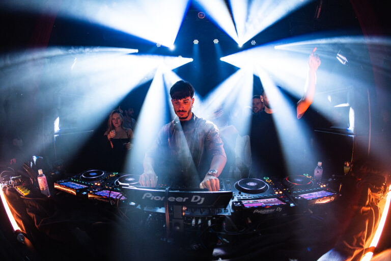 Ein Mann steht hinter einem Pioneer-DJ-Mischpult in einem Club unter hellen Bühnenscheinwerfern, während im Hintergrund eine Menschenmenge zu sehen ist.