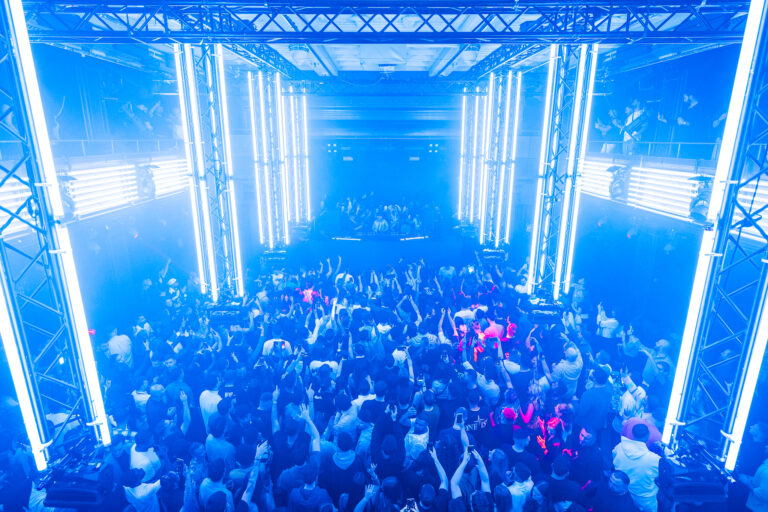 Ansicht von oben auf eine dichte Menschenmenge in einem großen Club in intensivem blauen Licht, umrahmt von vertikalen Leuchtröhren an Metalltraversen.
