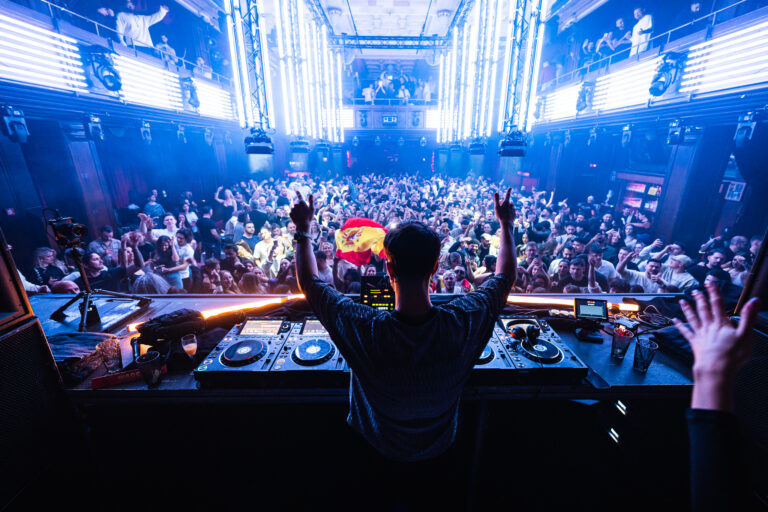 Ein Mann steht hinter einem DJ-Mischpult und hebt seine Arme vor einem großen, vollen Club unter blauem Bühnenlicht und vertikalen Leuchtröhren.