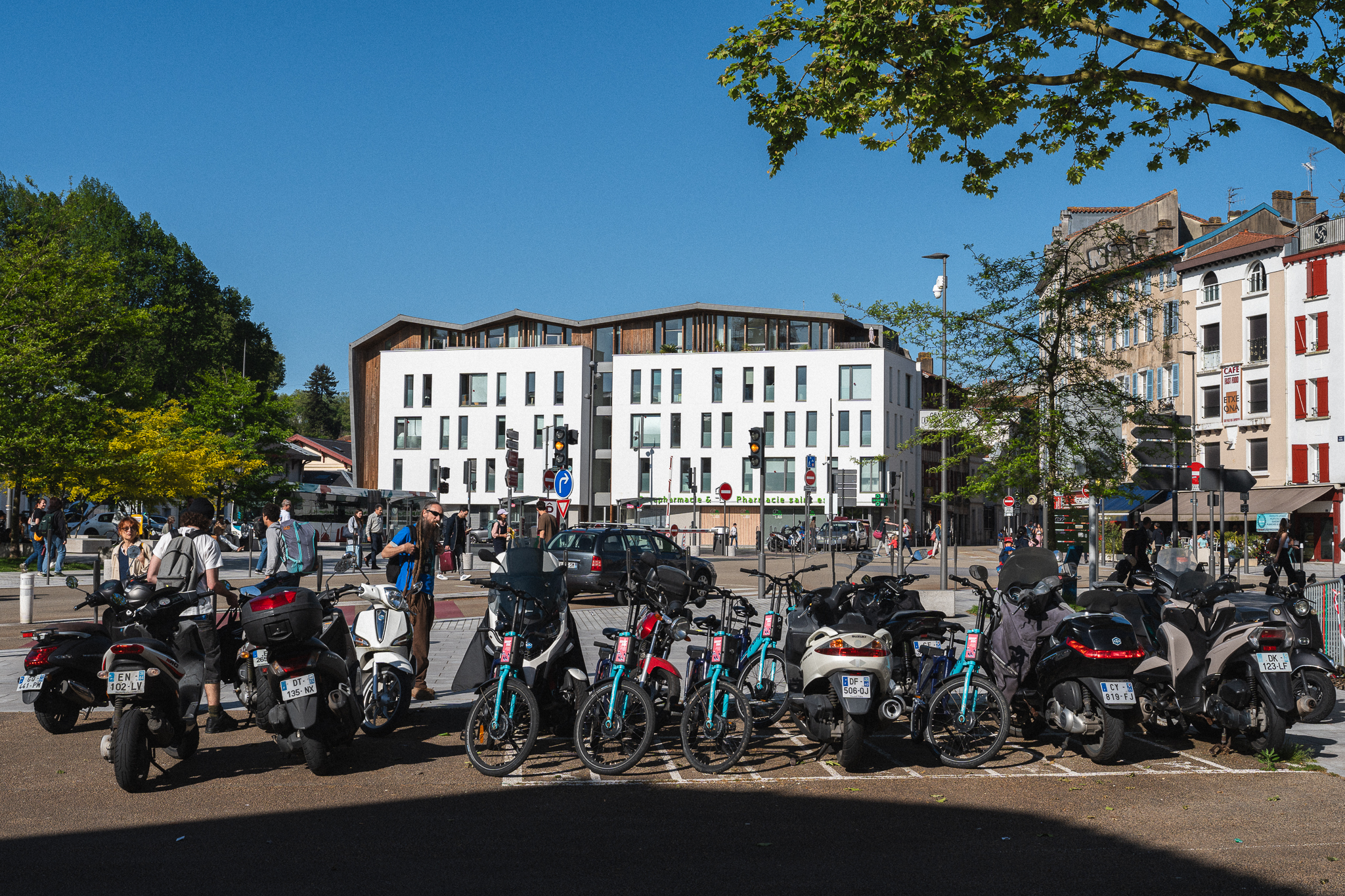 Egy sor parkoló motorkerékpár és kerékpár egy kövezett városi főtéren, a háttérben modern fehér épületekkel és emberekkel kék ég alatt.