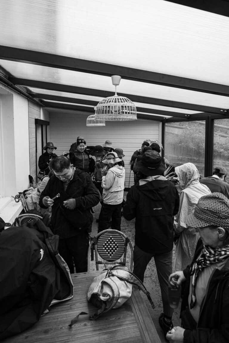 Fekete-fehér fényképen több kalapos és szabadtéri kabátos férfi és nő látható, amint egy üvegtetős télikertben gyülekeznek.