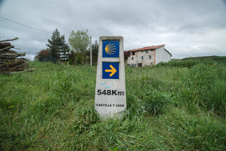 Egy betonból készült zarándokút-kilométerkő áll a zöld fűben borús ég alatt, sárga kagylóval, jobbra mutató nyílnak és az 548Km felirattal.