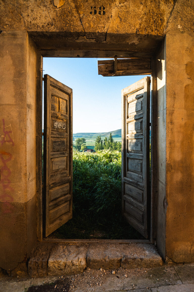 Open weathered wooden doors in a stone wall frame a view of green fields and rolling hills under a blue sky.
