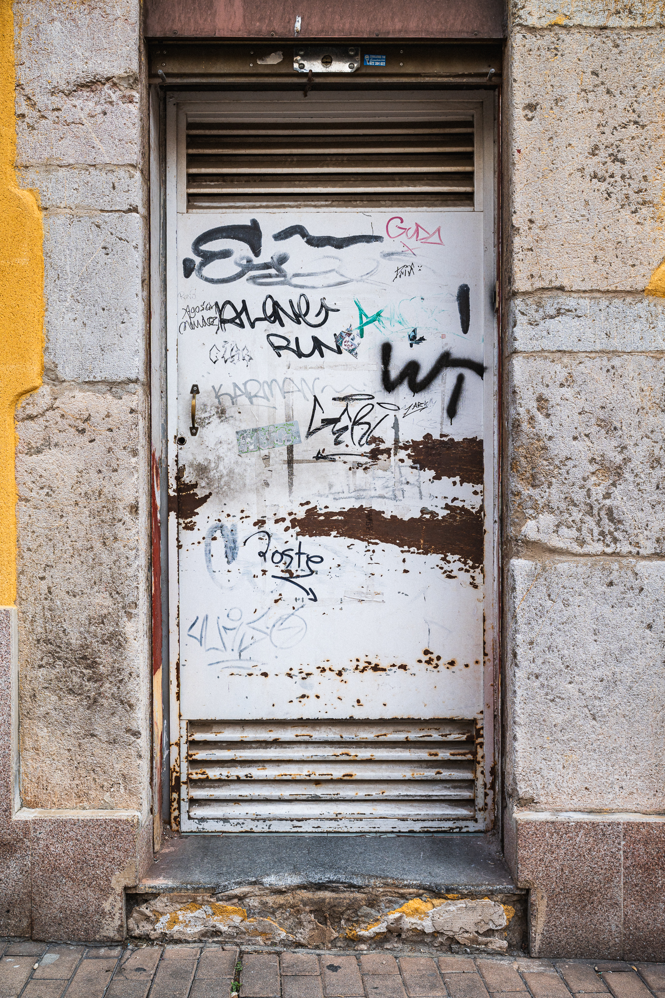 Graffitikkel és rozsdafoltokkal borított, viharvert fehér fémajtó texturált kőfalba ágyazva.