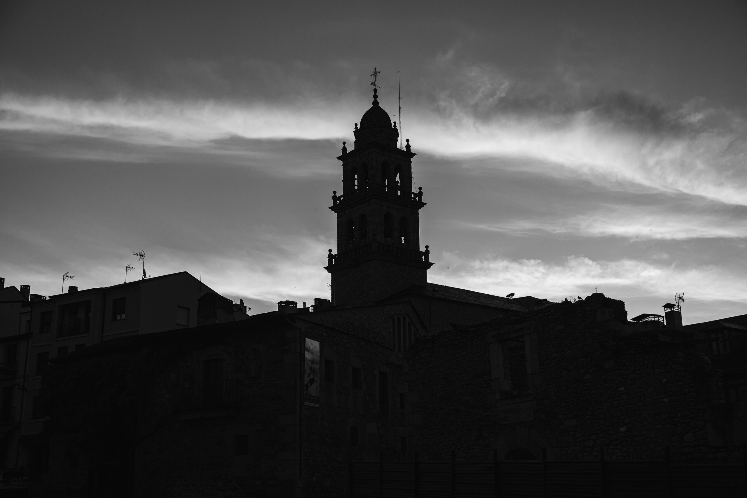 Templomtorony sziluettje felhős égbolt előtt, fekete-fehér fényképen.
