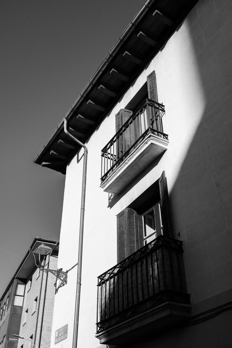 Low-angle black and white shot of a building facade with two balconies, illuminated by a diagonal beam of strong sunlight.
