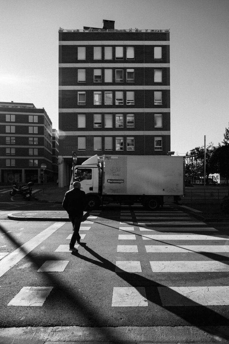A man walks on a pedestrian crosswalk in front of a white delivery truck and a tall apartment building, captured in a black-and-white street photograph.
