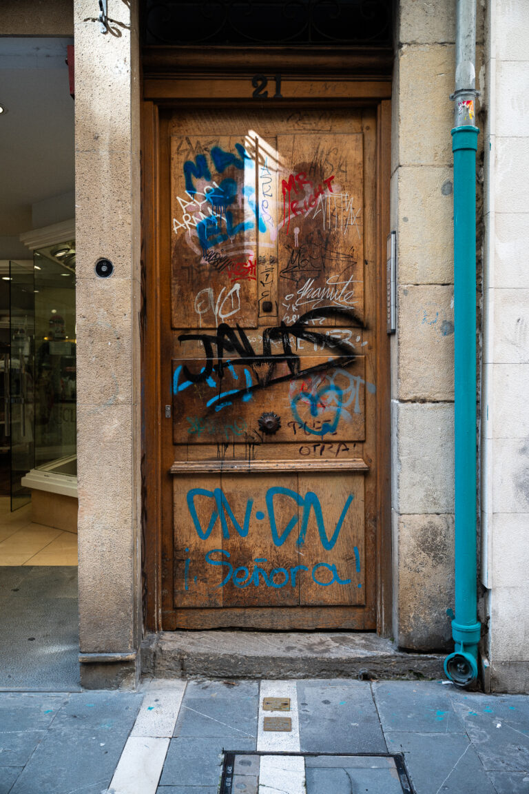 Egy viharvert, színes grafittikkel borított fa ajtó, felette a 21-es számmal, jobb oldalon egy élénktürkiz esővízcsatornával.