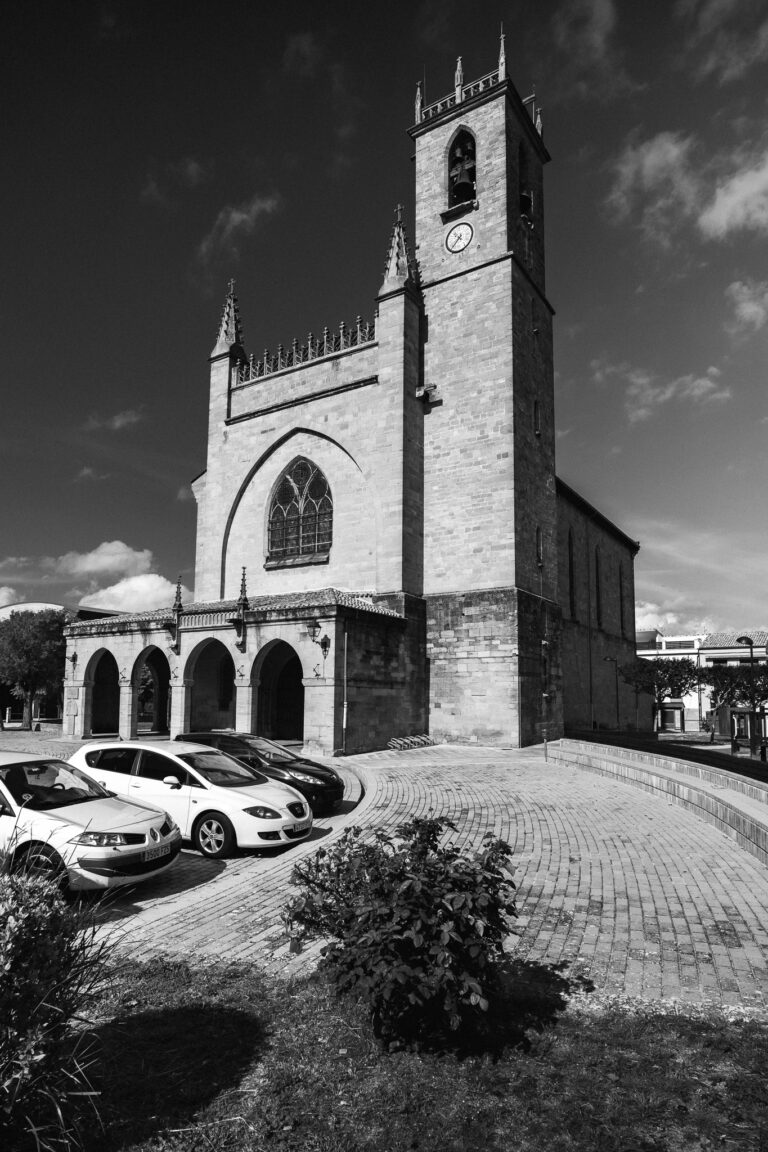 Fekete-fehér felvétel egy kőtemplomról harangtoronnyal, árkádos bejárattal és parkoló autókkal a kövezett téren.