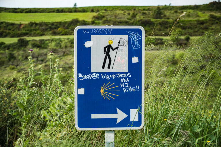 Grafittival borított kék Camino de Santiago tábla áll magas zöld fű és dombok között.