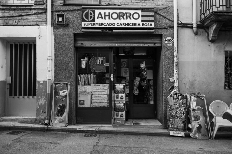 Fekete-fehér utcaszintű fényképen az Ahorro Supermercado Carniceria Rosa üzlethelyisége látható feliratokkal és kihelyezett állványokkal.