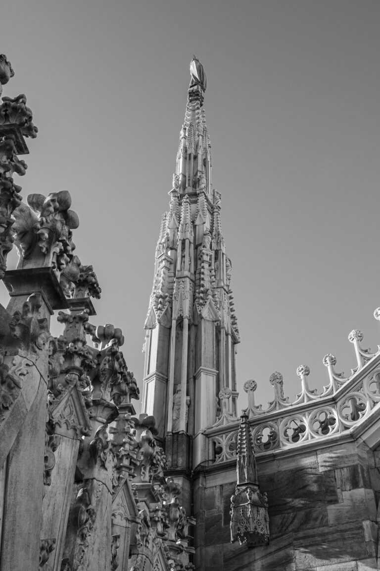 Eine Schwarz-Weiß-Ansicht aus niedriger Perspektive zeigt die gotische Turmspitze und die ornamentalen architektonischen Details des Mailänder Doms vor einem klaren Himmel.