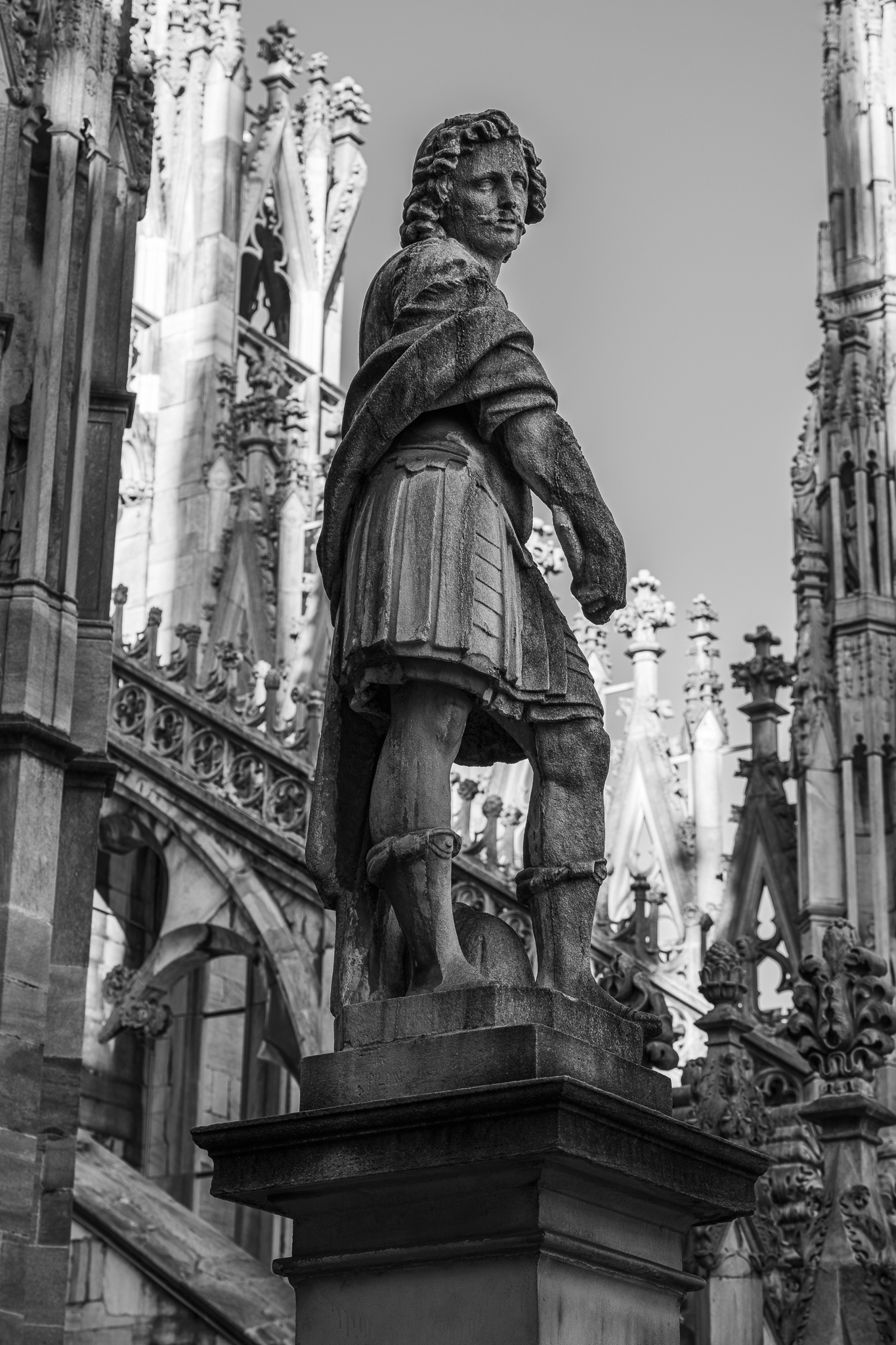 Eine Schwarz-Weiß-Ansicht aus niedriger Perspektive zeigt die steinerne Statue eines Mannes auf einem Sockel vor einer reich verziertem Kathedralenarchitektur.