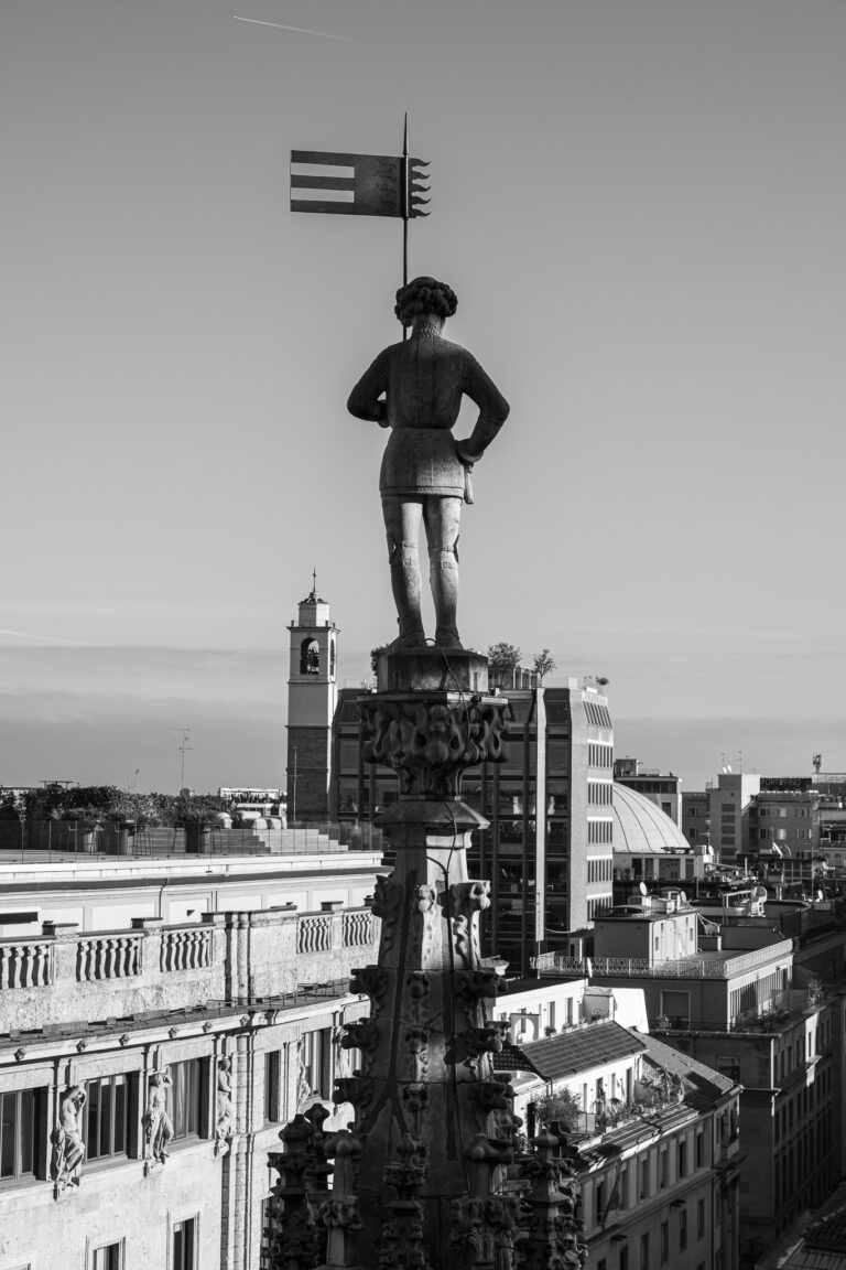 Eine Schwarz-Weiß-Aufnahme zeigt die Statue eines Mannes mit einer Flagge auf einer verzierten Fiale, mit Blick auf Dächer der Stadt.