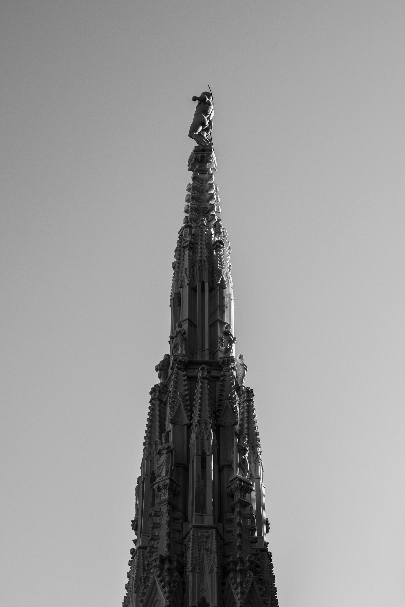 Eine schwarz-weiße Untersicht einer prunkvollen gotischen Kathedralenspitze mit einer kleinen Figur vor klarem Himmel.