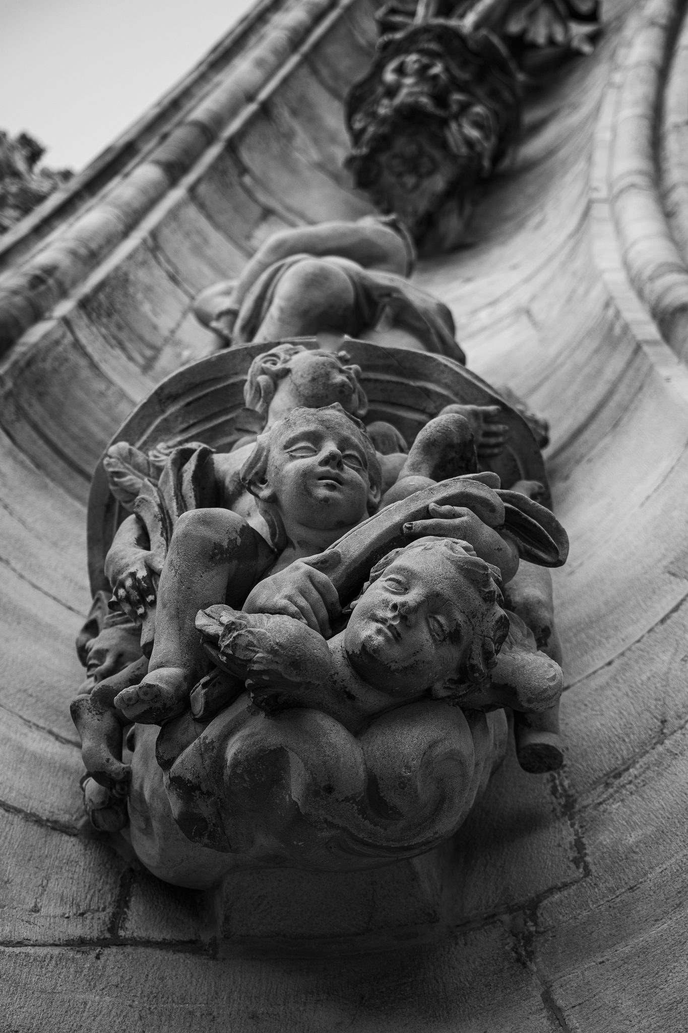 Schwarz-Weiß-Fotografie aus niedriger Perspektive zeigt detaillierte Steinmetzarbeiten von Figuren und Putten an einer verzierten Architekturfassade.