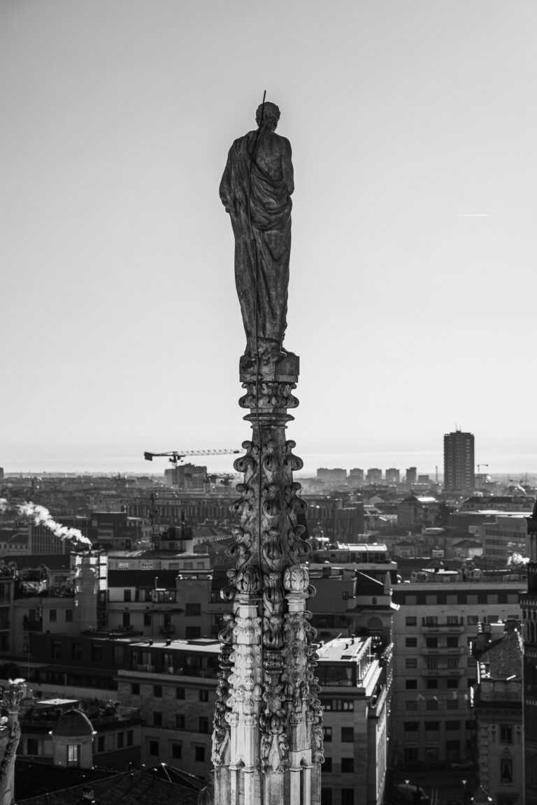 Eine Schwarz-Weiß-Ansicht von oben zeigt die Statue eines Mannes auf einer verzierten architektonischen Fiale mit Blick auf eine entfernte Stadtsilhouette.