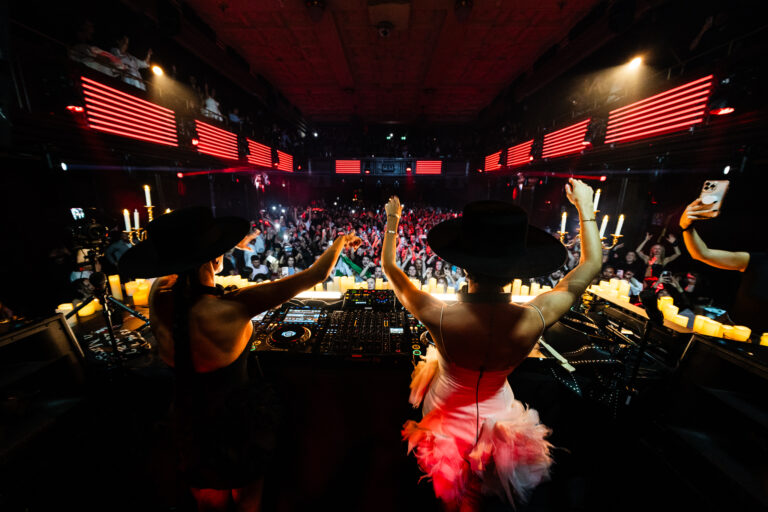 Two women wearing black hats stand behind a DJ console, raising their arms before a large crowd in a dark venue lit by candles and red lights.
