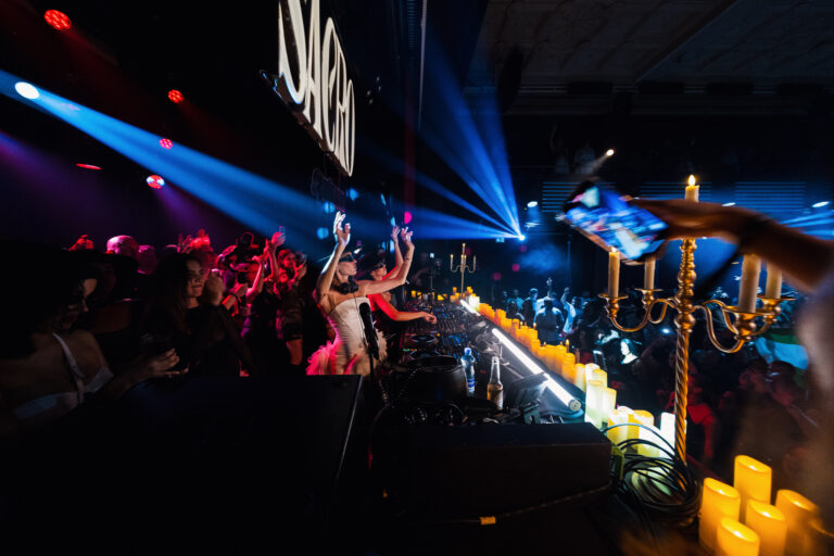 A woman stands behind a DJ booth under blue stage lights, facing a crowded club with candles lining the booth foreground.