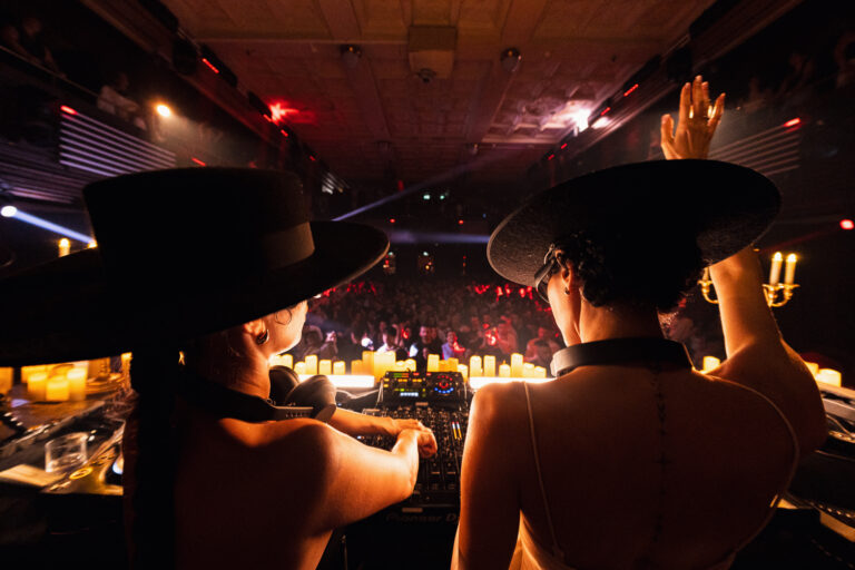 Two women wearing wide-brimmed hats stand behind a DJ mixer in a dimly lit club, facing a crowded dance floor illuminated by candles.