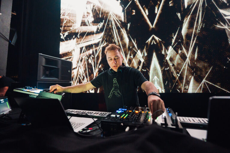 A man stands behind audio mixing equipment, operating a controller with his hands in a brightly lit indoor venue.