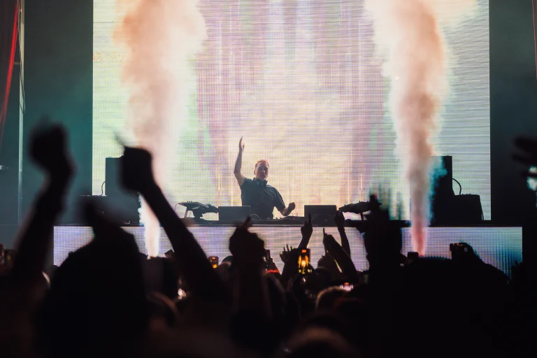 A man stands behind a DJ booth on stage raising his hand, framed by large vertical plumes of smoke and silhouetted concertgoers in the foreground.